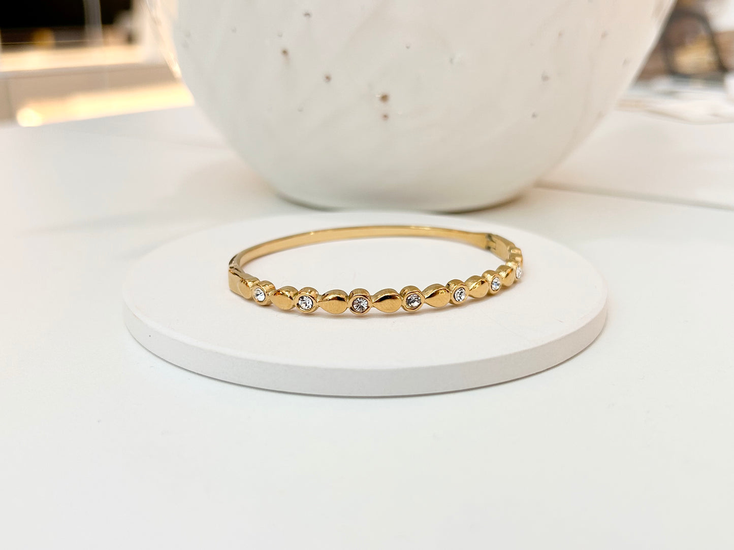 
                  
                    A delicate gold bangle bracelet with a row of small clear stones, displayed on a round white coaster with a white speckled ceramic vase in the background.
                  
                