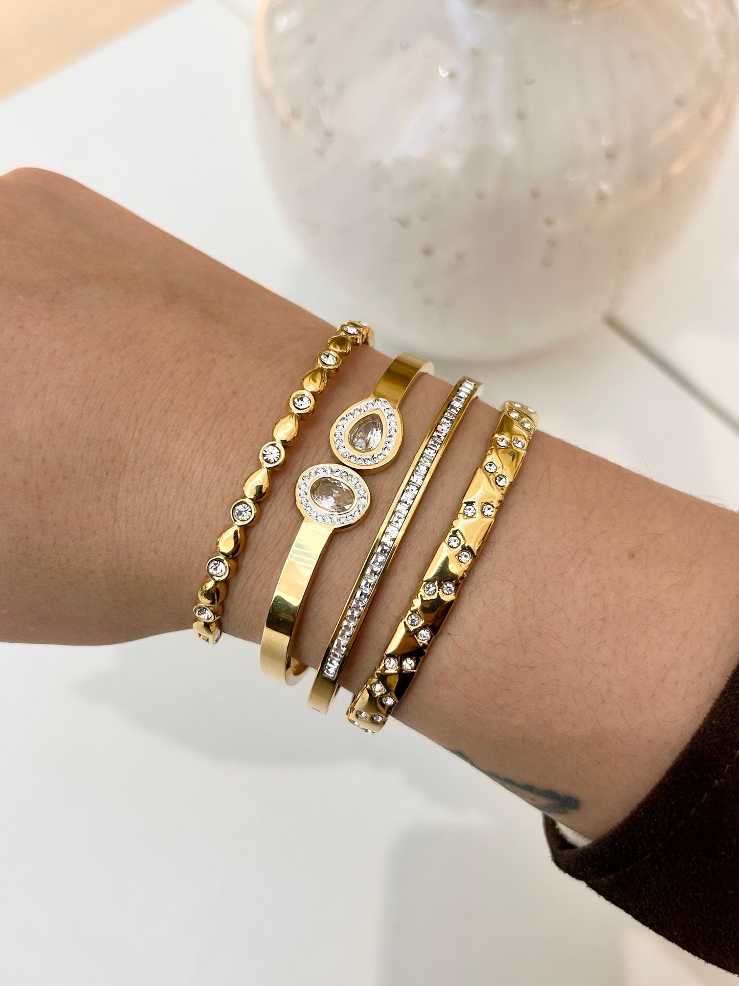 A close-up of a person's wrist wearing five stacked gold-toned bangles, including two with clear crystal details, against a soft white background.