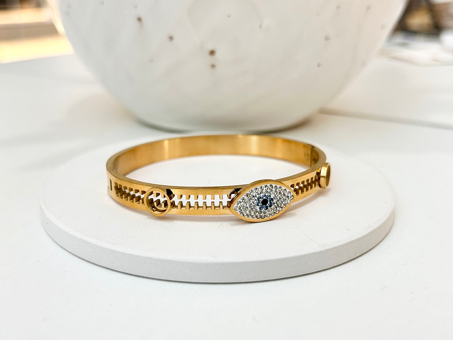 
                  
                    A gold bangle bracelet with a central evil eye motif paved with small clear stones and a dark blue center stone, displayed on a round white stand.
                  
                