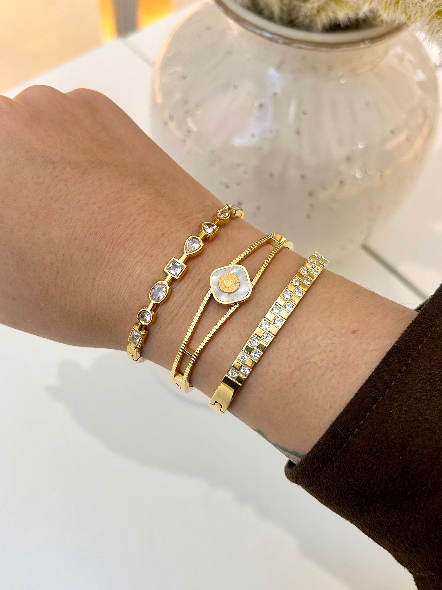 A close-up of a person's wrist wearing four gold bracelets, including a bezel-set crystal bracelet, a rope-style bracelet with a central oval charm, a cable-wrapped bracelet, and a crystal-encrusted bangle, with a white ceramic vase blurred in the background.