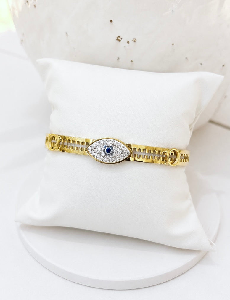 A gold-toned bracelet displayed on a white cushion stand. The bracelet features a central evil eye motif with a blue center stone and small clear stones around it, set against a softly blurred white background.