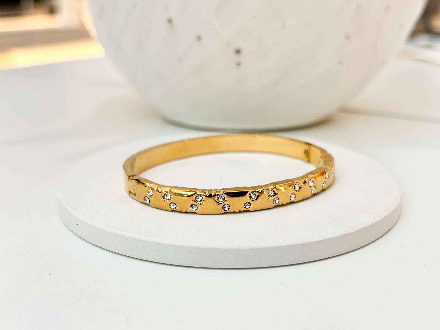 
                  
                    A gold bangle bracelet with small clear rhinestones set along the front, displayed on a round white coaster with a white ceramic bowl in the background.
                  
                