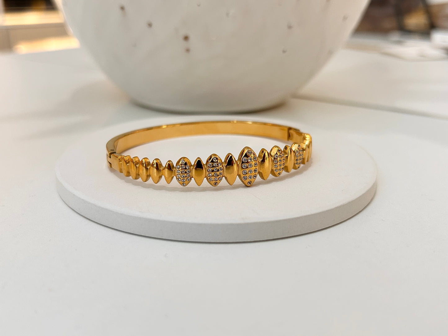 
                  
                    A gold bangle bracelet with a row of decorative pavé and solid teardrop-shaped links, displayed on a round white stand.
                  
                
