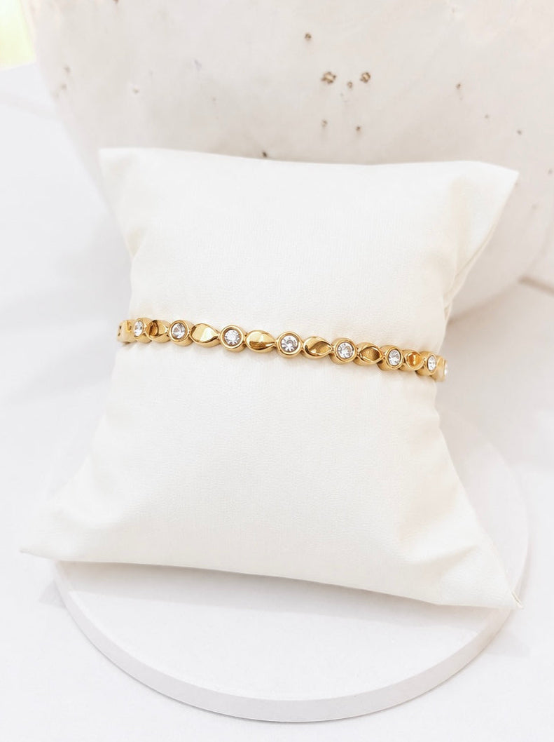 A delicate gold bracelet with small clear stones, displayed on a white satin pillow against a white background with a decorative ceramic vase.
