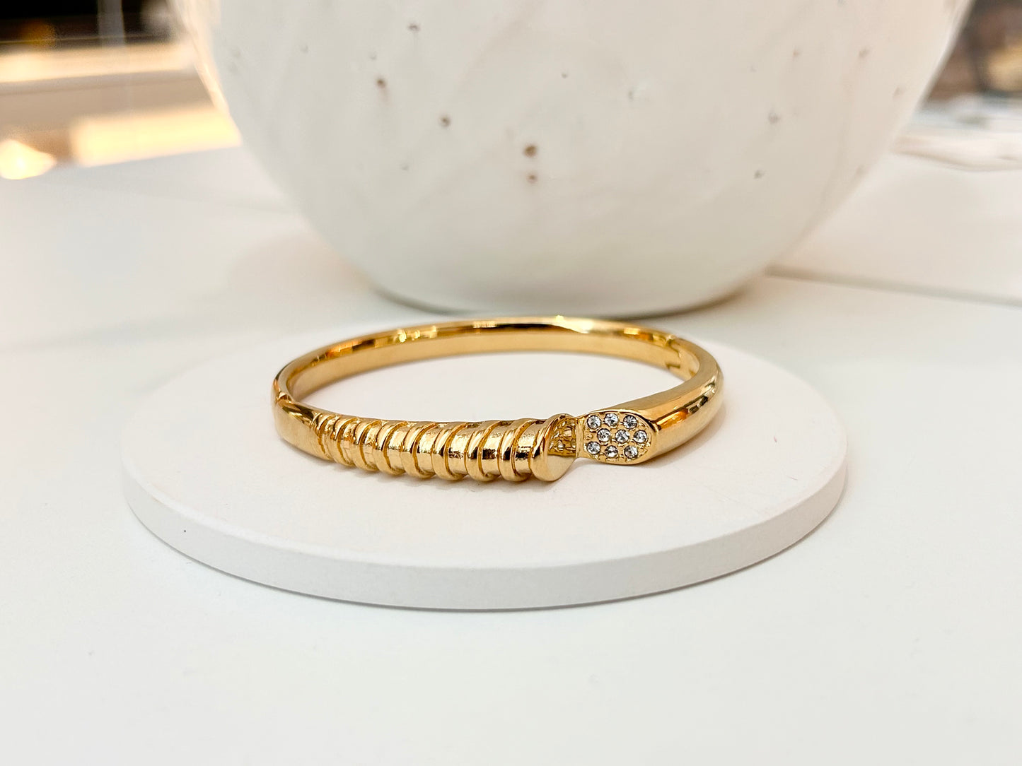 
                  
                    A gold bangle bracelet with a textured ridged band and a small pavé-set clear stone accent, displayed on a round white pedestal.
                  
                