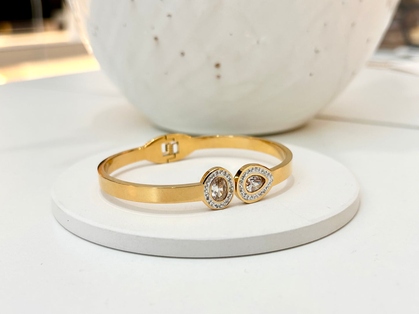 
                  
                    A gold bangle bracelet with two oval bezel-set clear stones, displayed on a round white pedestal with a white speckled ceramic vase in the background.
                  
                
