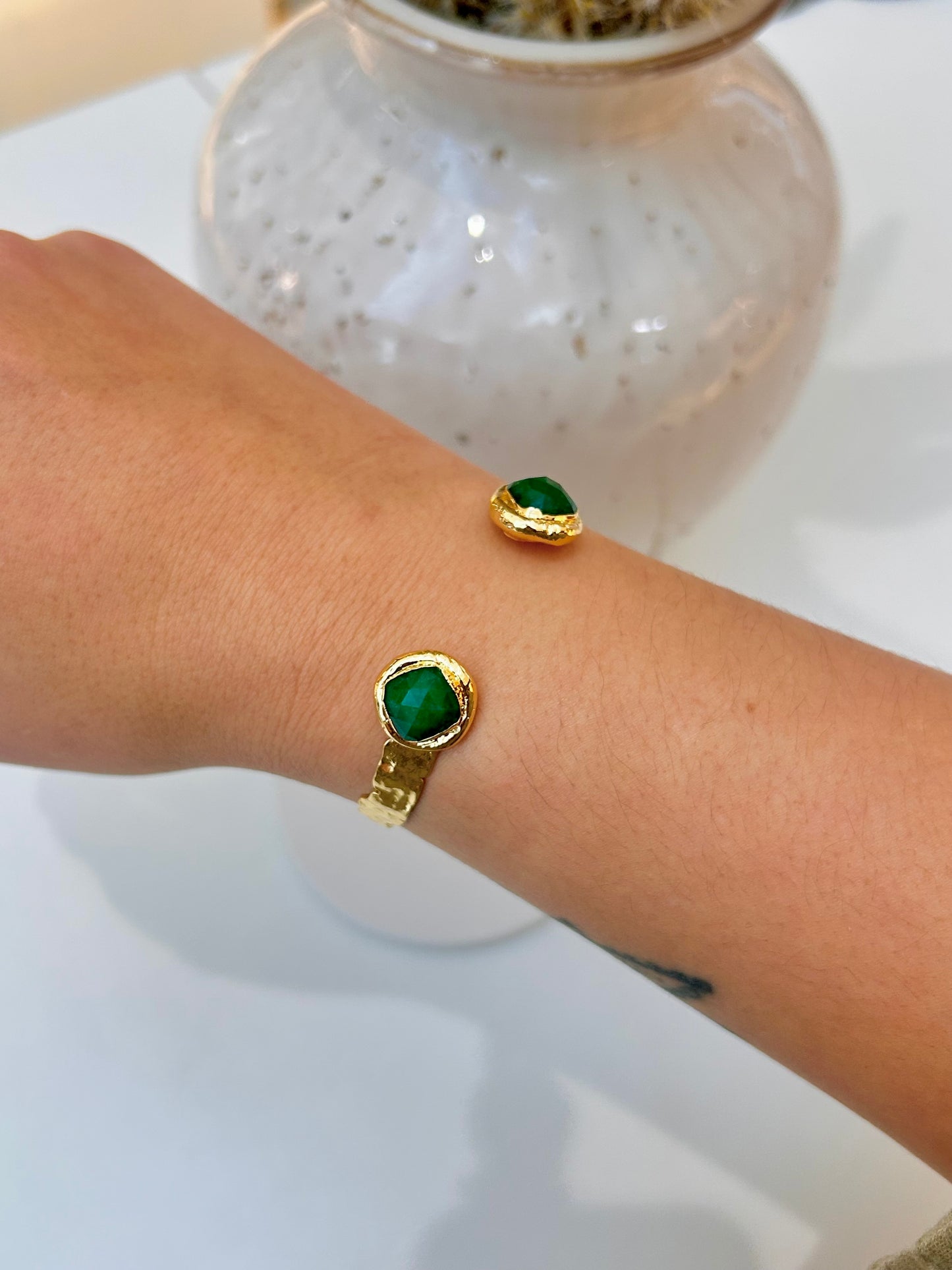 
                  
                    A close-up of a person's wrist wearing two gold bracelets, each featuring a round green cabochon stone, with a white speckled vase blurred in the background.
                  
                