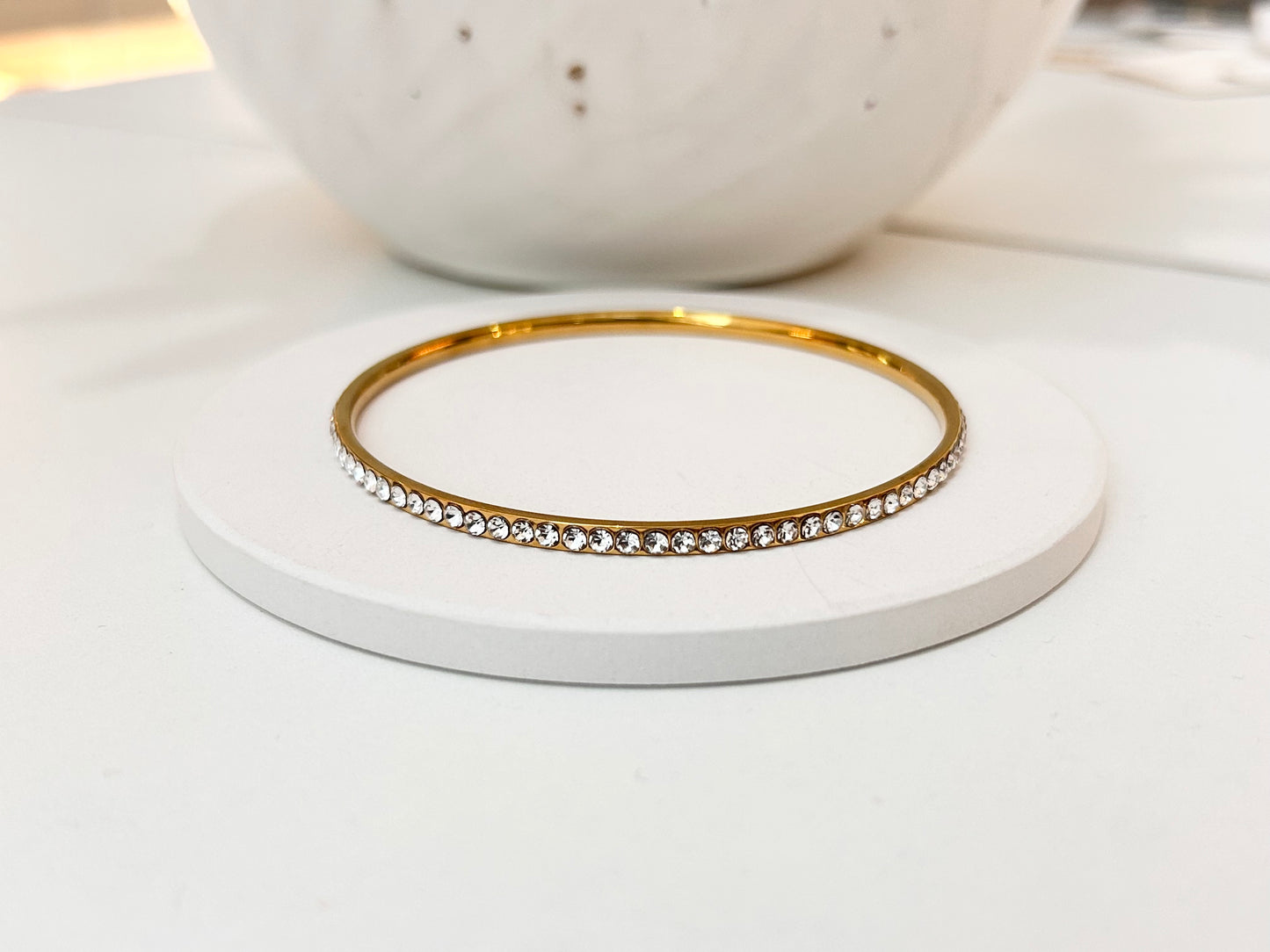 
                  
                    A thin gold bangle bracelet with a row of clear rhinestones, displayed on a round white coaster.
                  
                