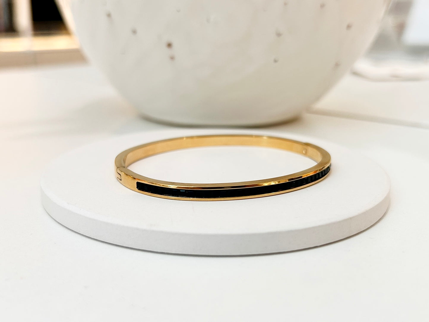 
                  
                    A slim gold bangle bracelet with a black inlay, displayed on a round white coaster with a speckled white ceramic bowl in the blurred background.
                  
                