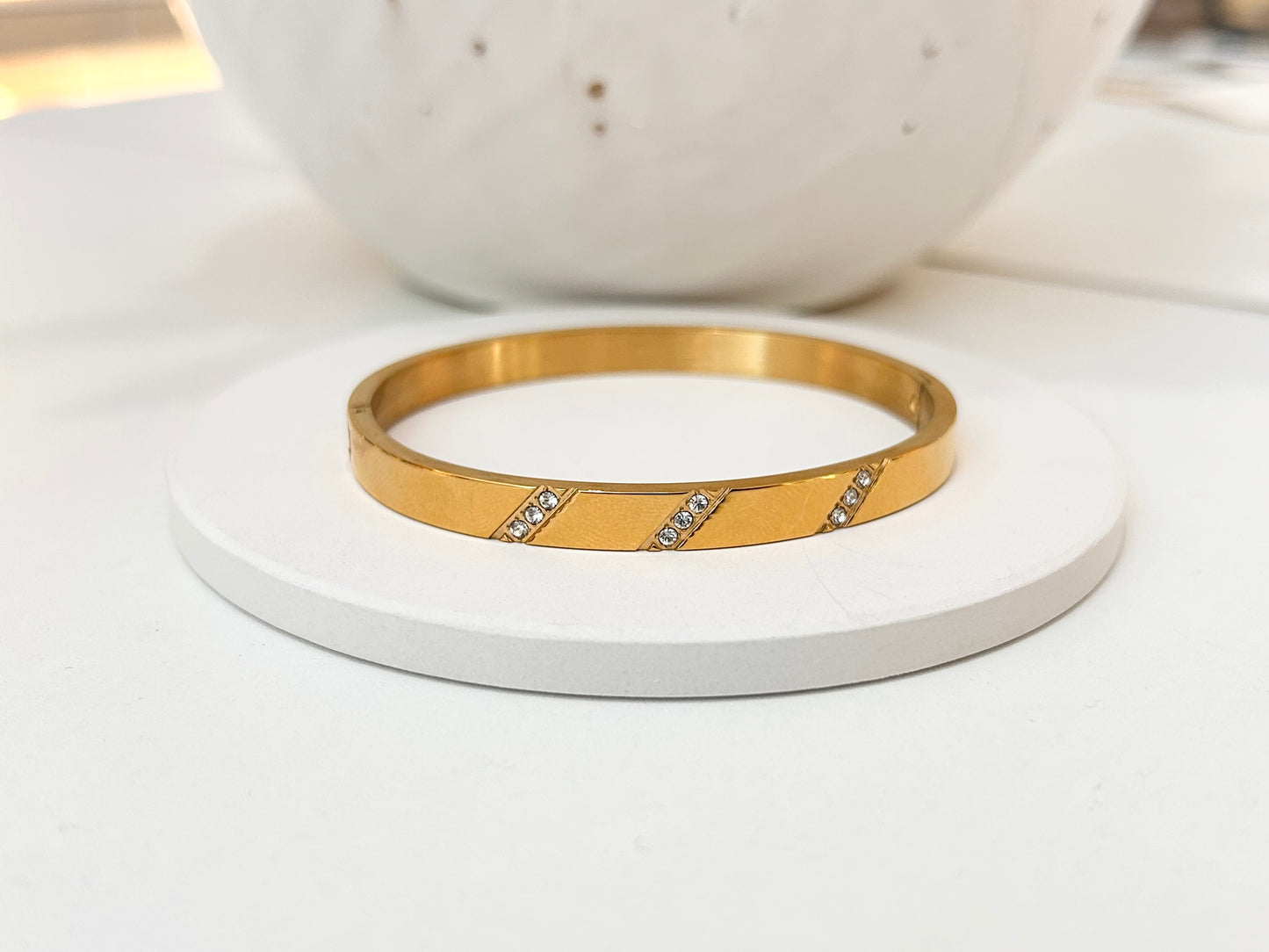 
                  
                    A gold bangle bracelet with two diagonal rows of small clear stones, displayed on a round white coaster with a white ceramic bowl in the background.
                  
                