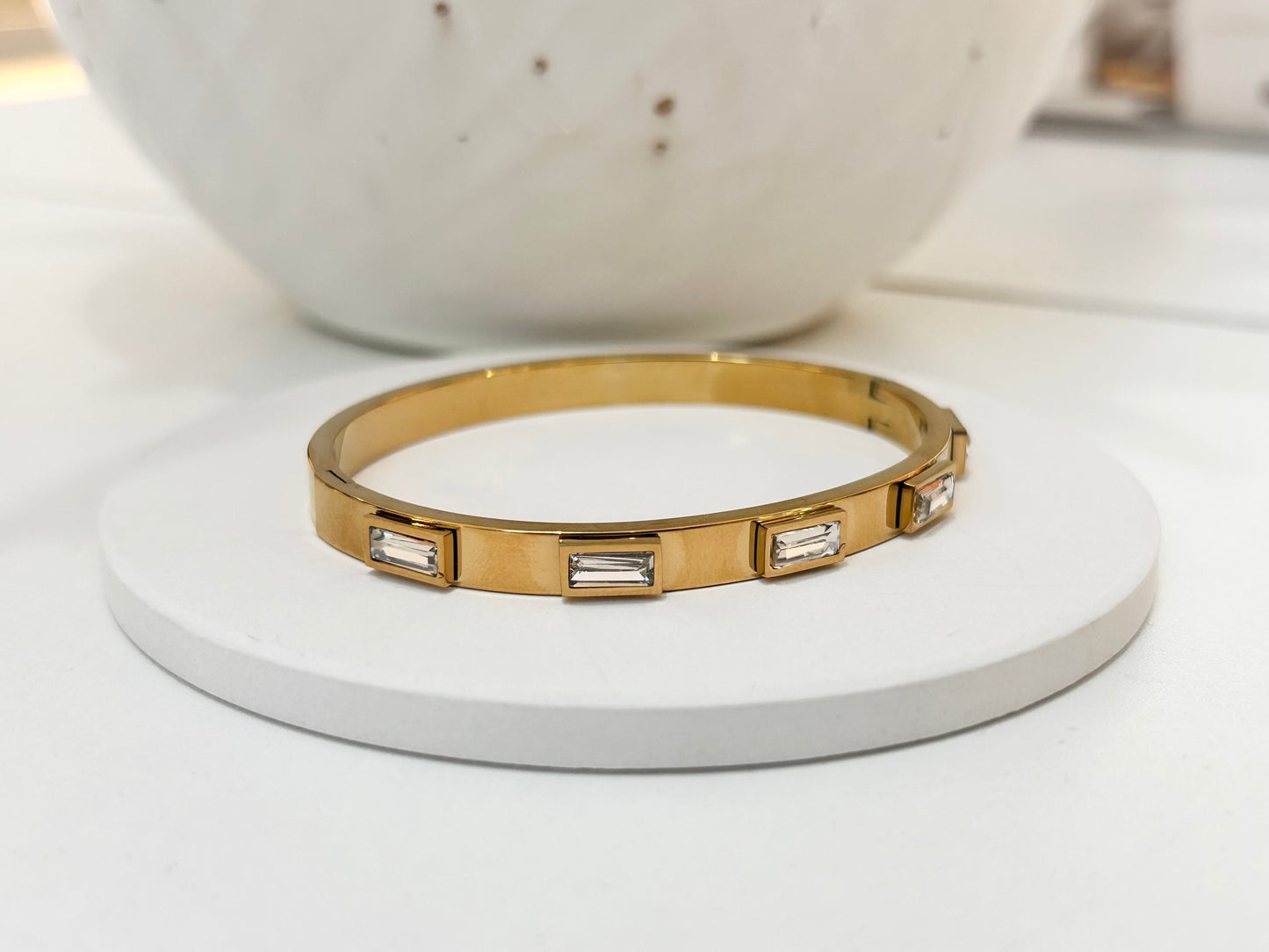
                  
                    A gold bangle bracelet with rectangular baguette-cut clear stones, displayed on a round white coaster with a white ceramic bowl in the background.
                  
                