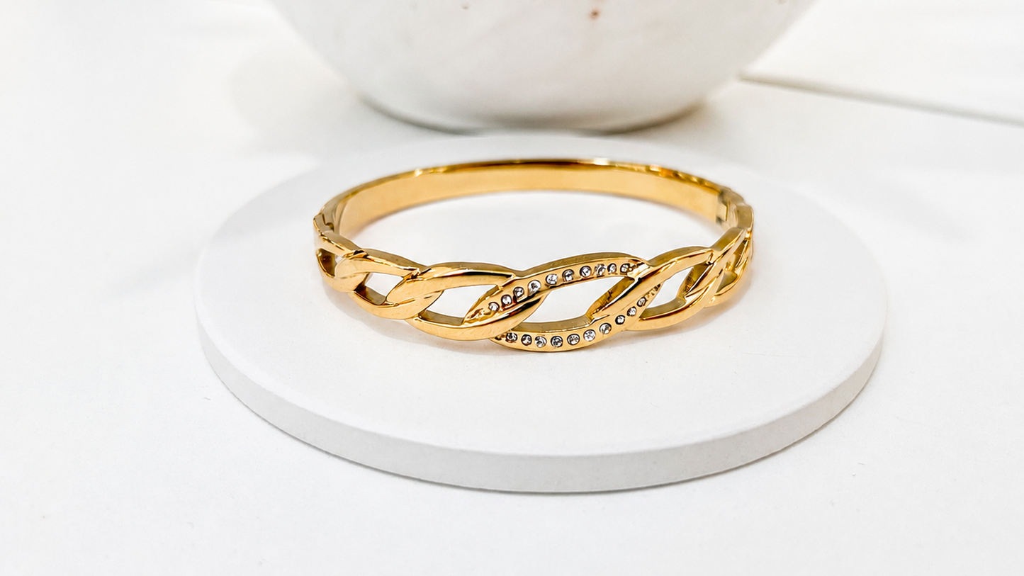 A gold infinity-style bangle bracelet with small clear stones set along the center, displayed on a round white coaster with a white ceramic bowl in the blurred background.