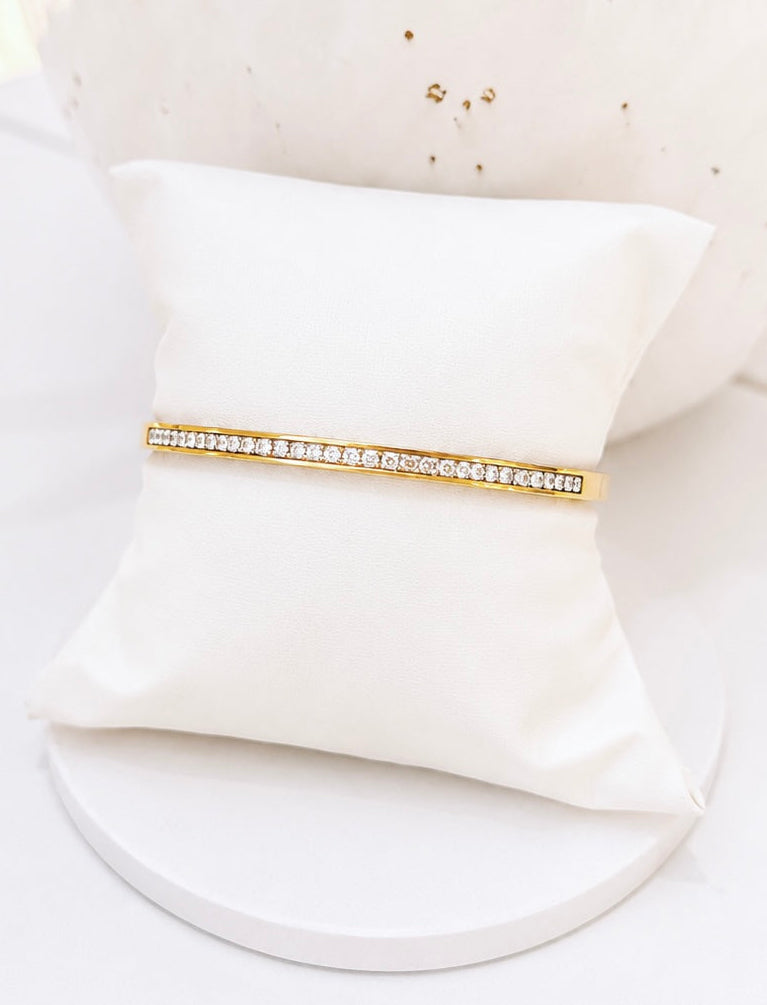 A gold bangle bracelet with a row of clear stones, displayed on a small white pillow against a white background.