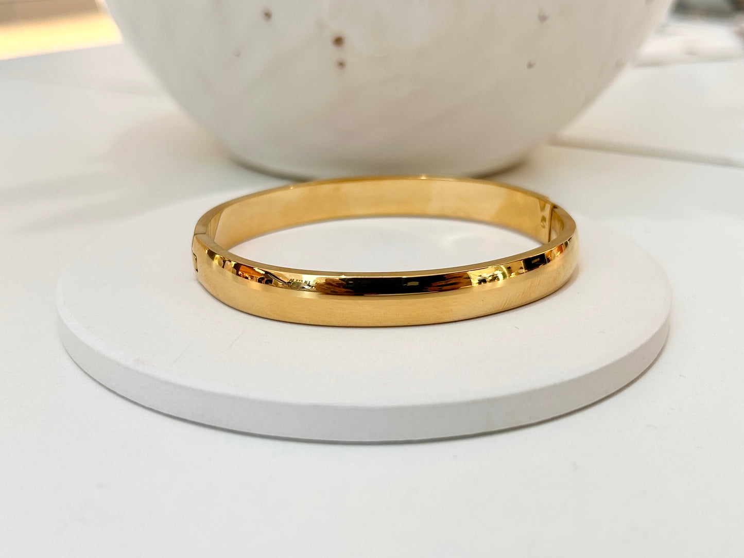 
                  
                    A polished gold bangle bracelet displayed on a round white coaster, with a blurred white ceramic bowl in the background.
                  
                