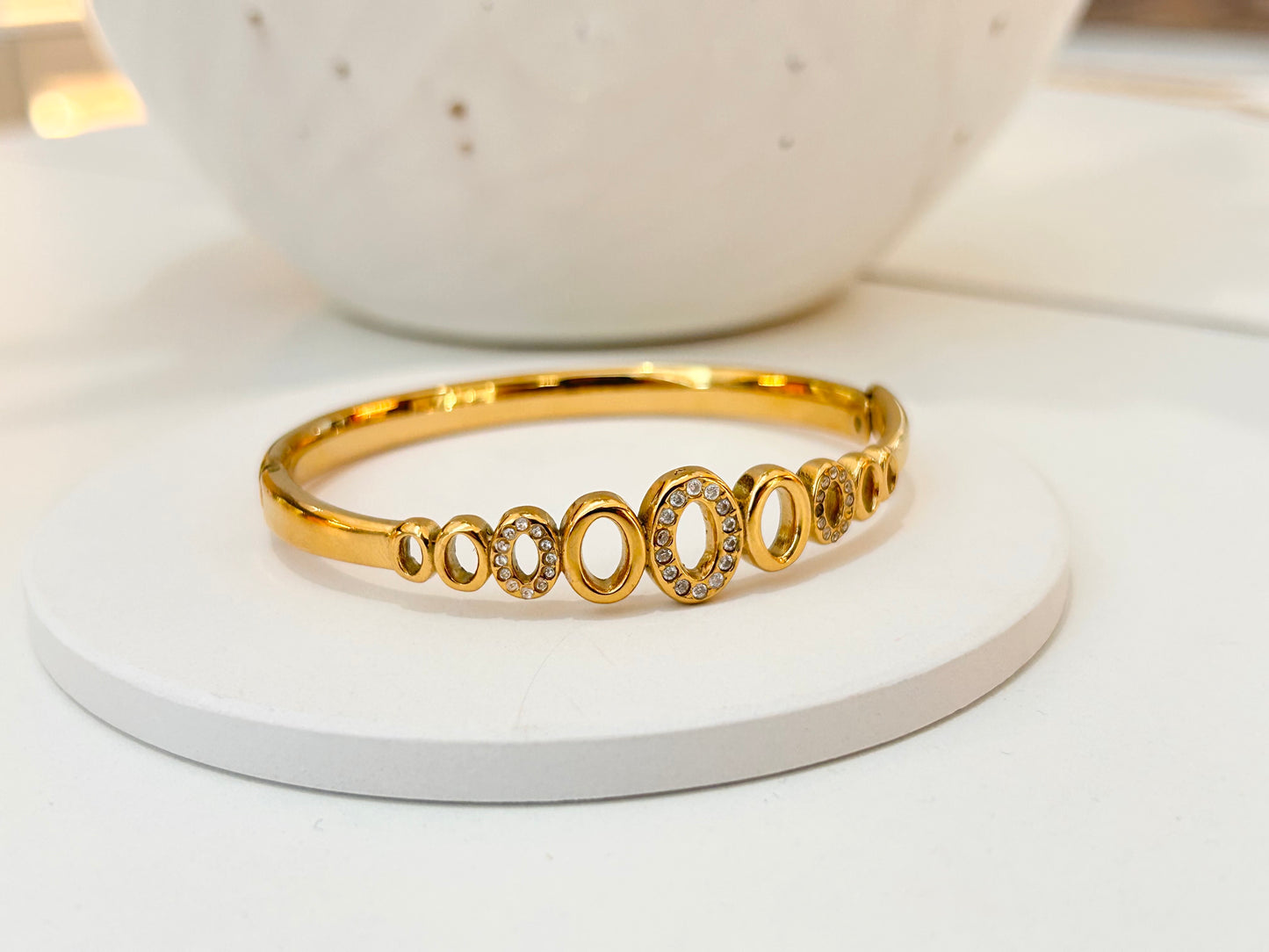 
                  
                    A gold bangle bracelet with a row of oval links, some featuring clear pavé stones, displayed on a white round pedestal.
                  
                