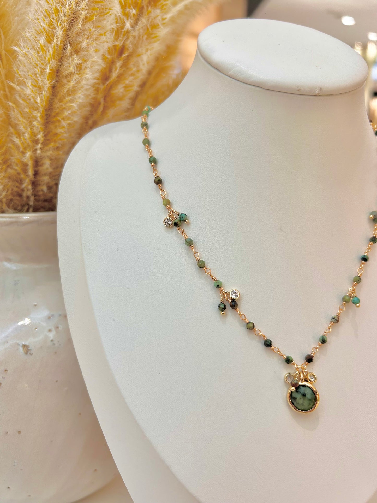 
                  
                    A delicate gold beaded necklace with small African turquoise beads and a green natural stone pendant, displayed on a white jewelry bust.
                  
                
