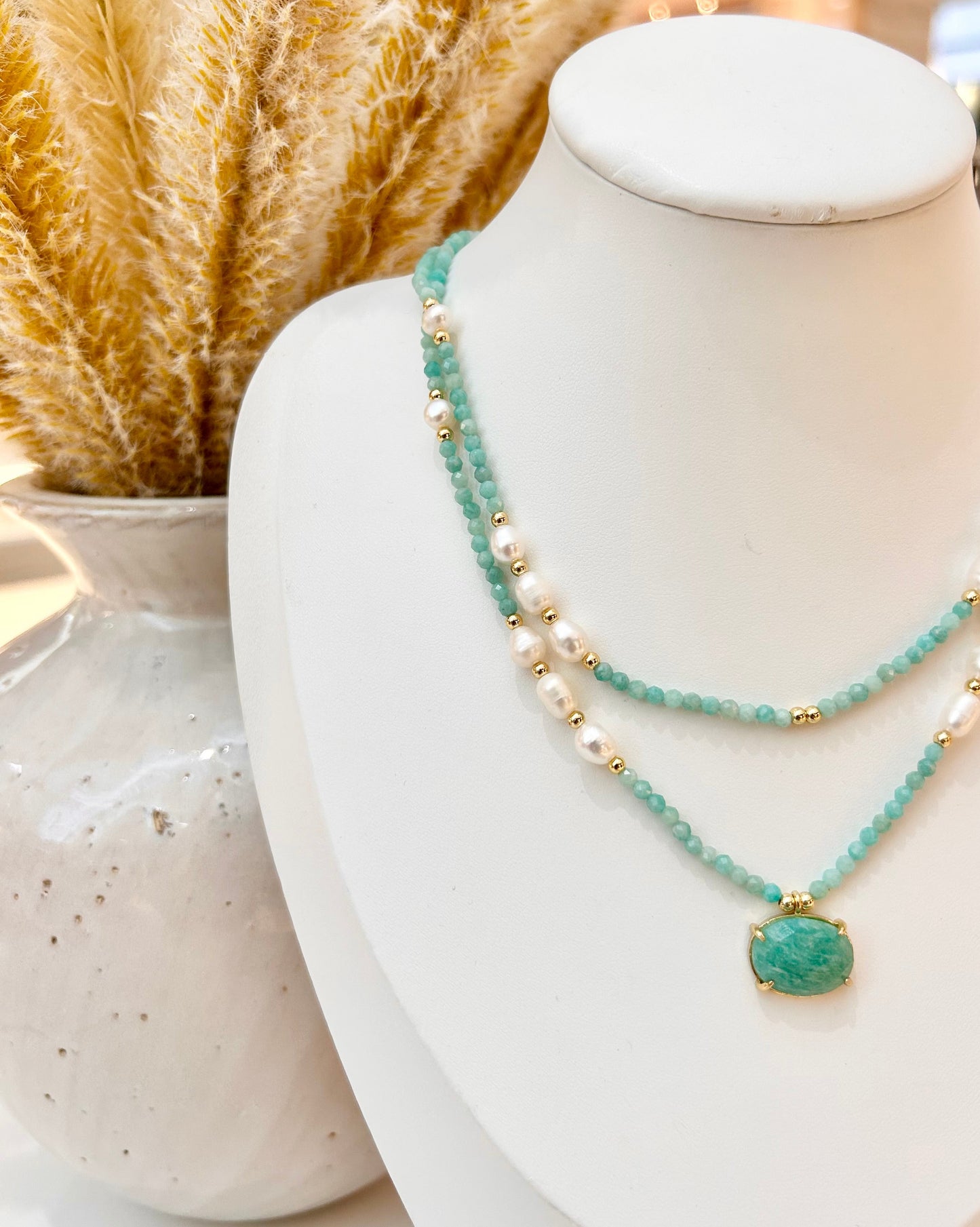A double-strand necklace displayed on a white mannequin bust, featuring a strand of amazonite faceted beads and a strand of white pearls with small gold spacer beads. The necklace has a large oval light green stone pendant in a gold bezel setting.
