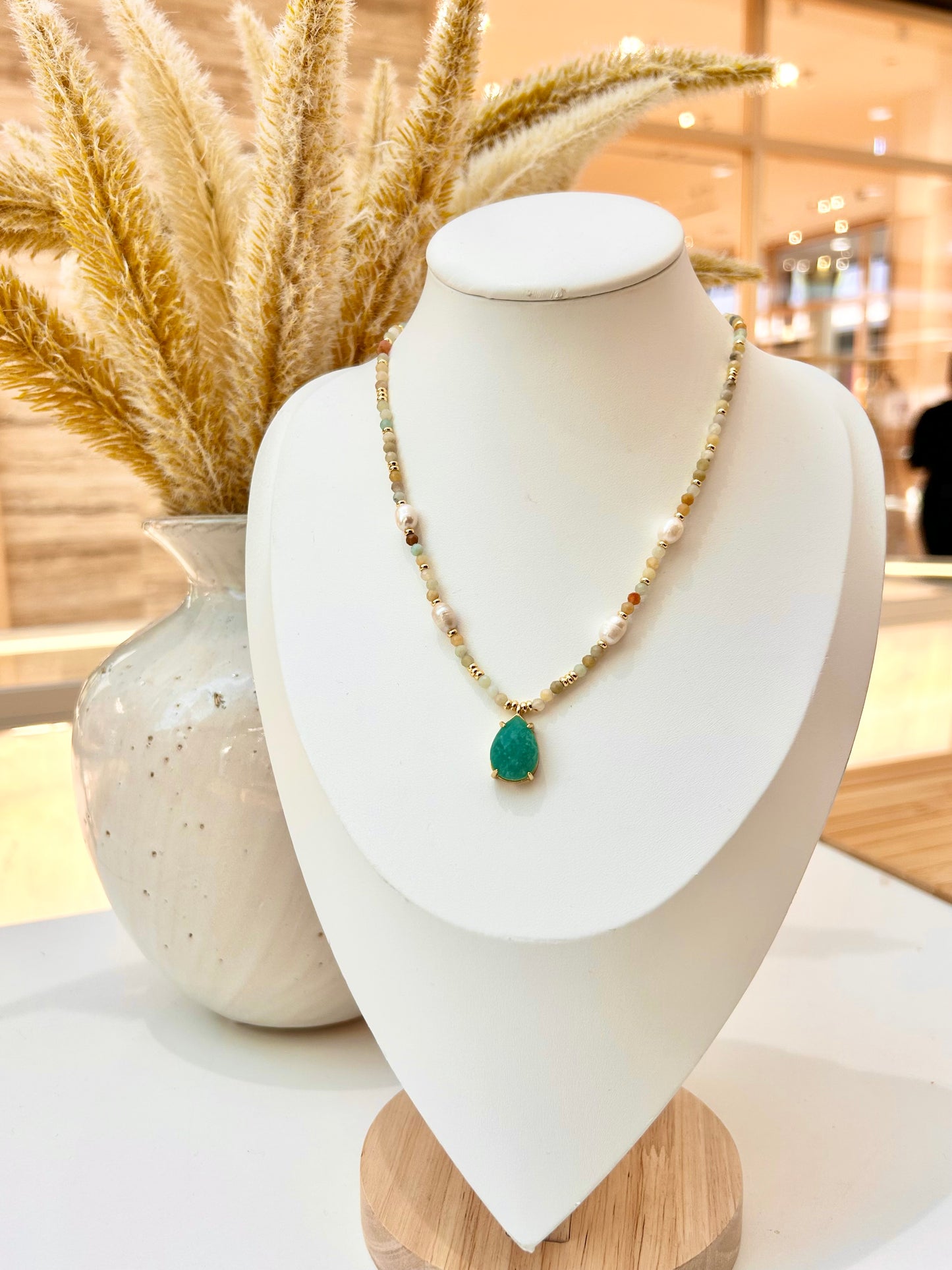
                  
                    A gold beaded necklace with a amazonite teardrop pendant displayed on a white jewellery bust, with a vase of dried grass in the background.
                  
                