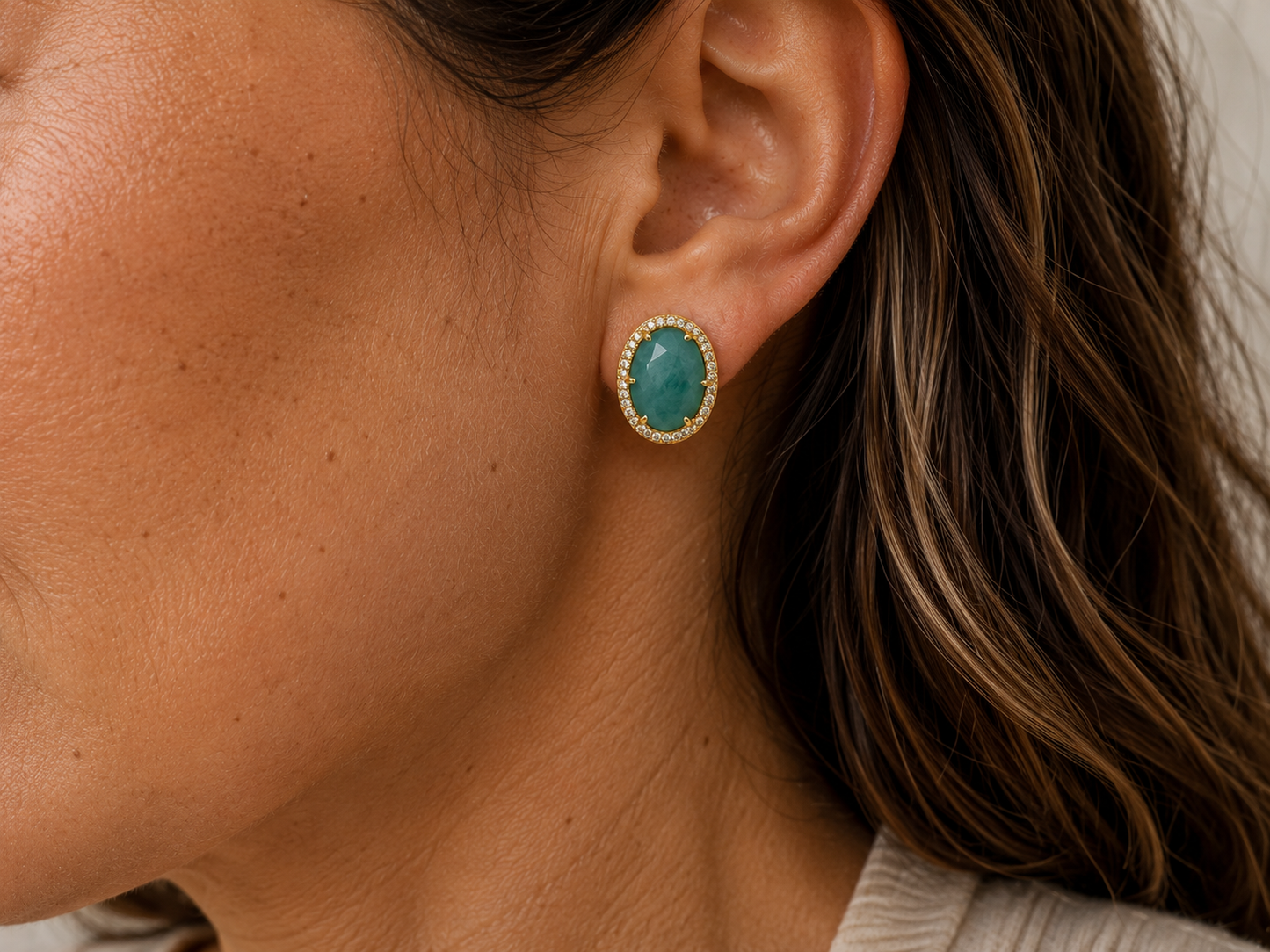 A close-up of a woman's ear wearing a gold stud earring with an oval amazonite stone surrounded by a textured gold border.