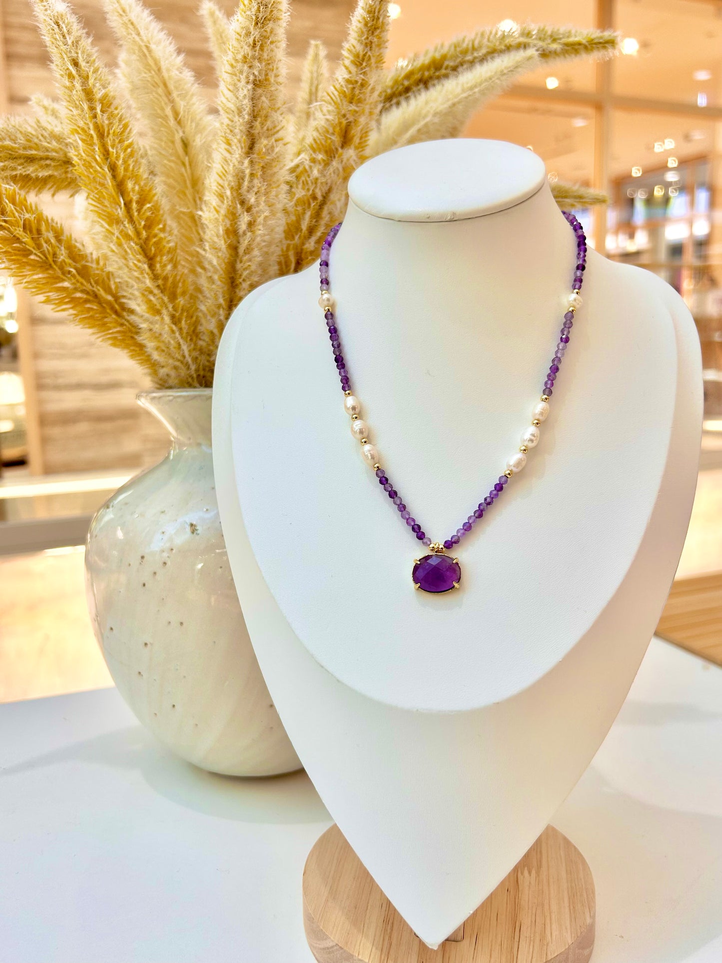 
                  
                    A beaded necklace with amethyst and freshwater pearl beads and an amethyst oval pendant, displayed on a white jewellery bust. A ceramic vase with dried pampas grass sits behind the bust in a softly lit indoor setting.
                  
                