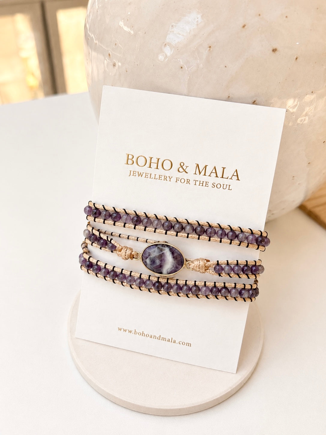 A stack of purple amethyst beaded bracelets with a central amethyst stone and gold-toned accents, displayed on a white card labeled BOHO & MALA, set against a soft beige background.