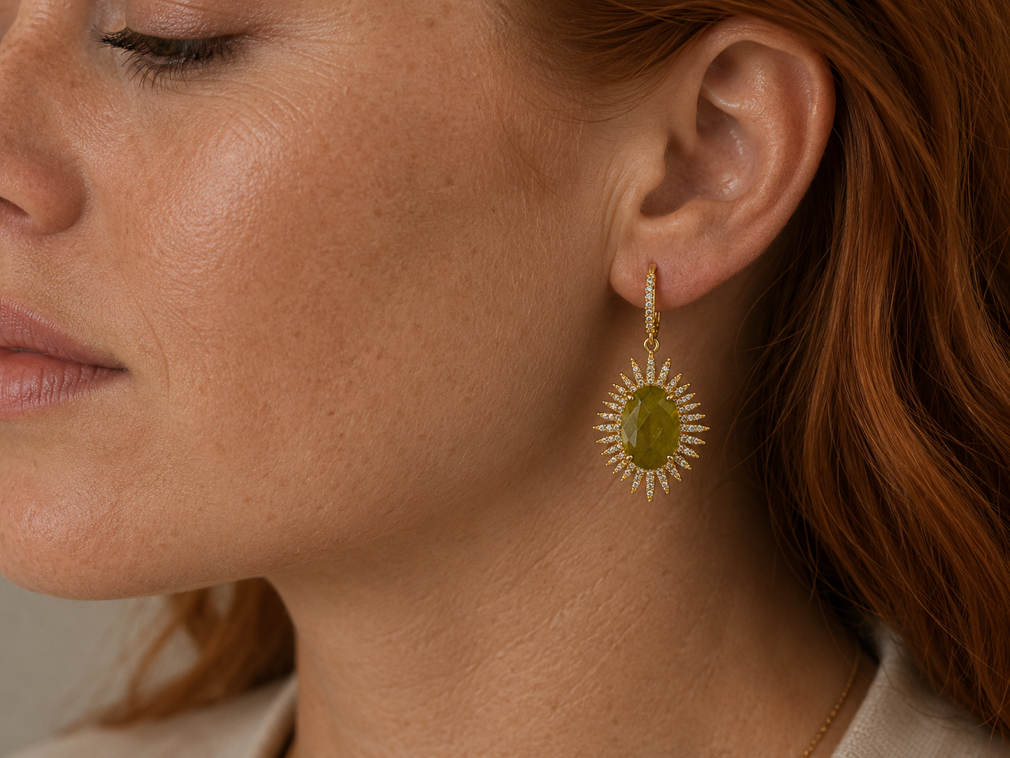 A close-up of a woman's ear wearing a gold dangle earring with a round olive-green center stone and a surrounding sunburst of small clear stones.