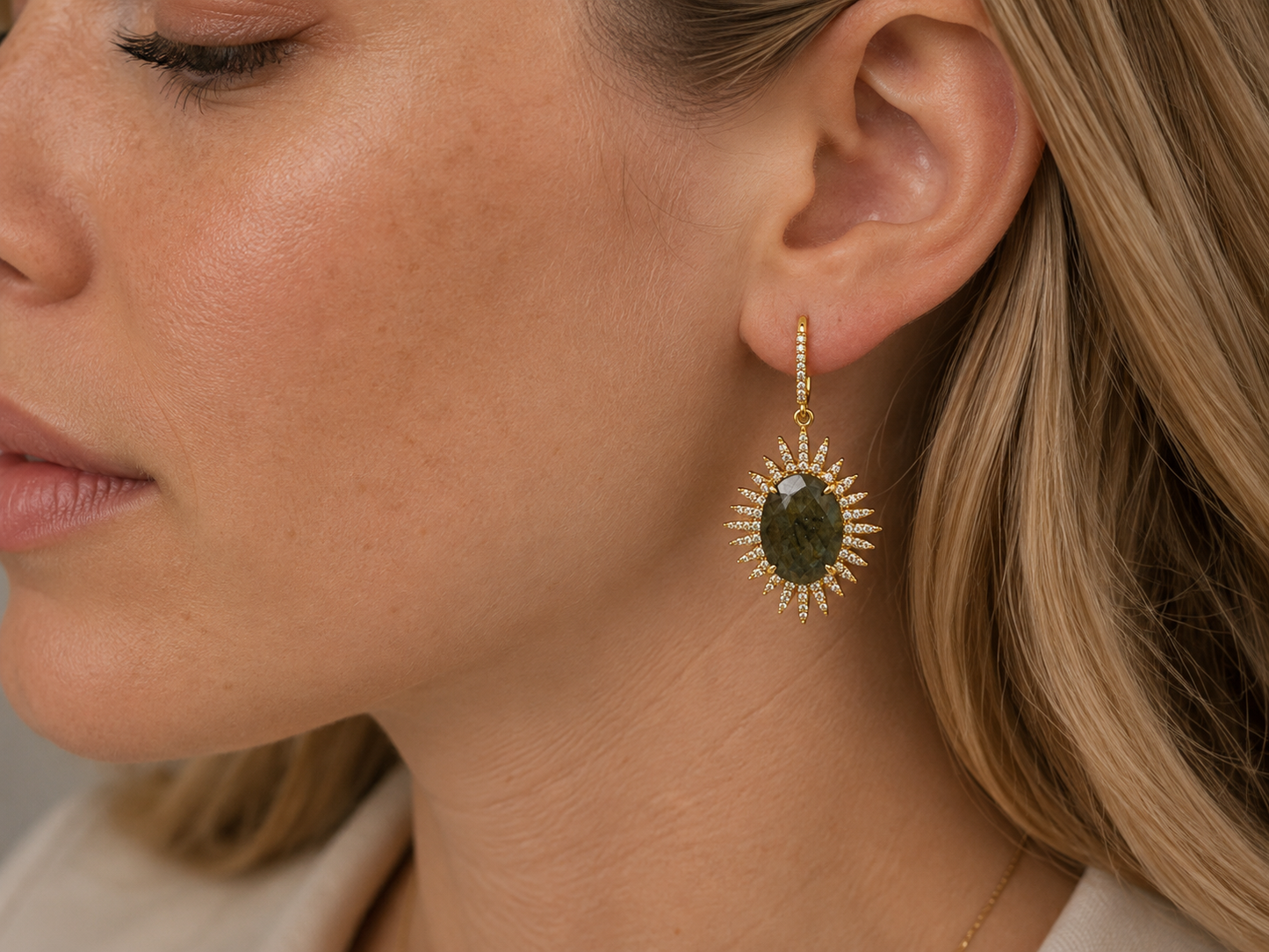 A close-up side profile of a woman wearing a gold drop earring with a round dark green stone surrounded by small clear stones in a sunburst setting.