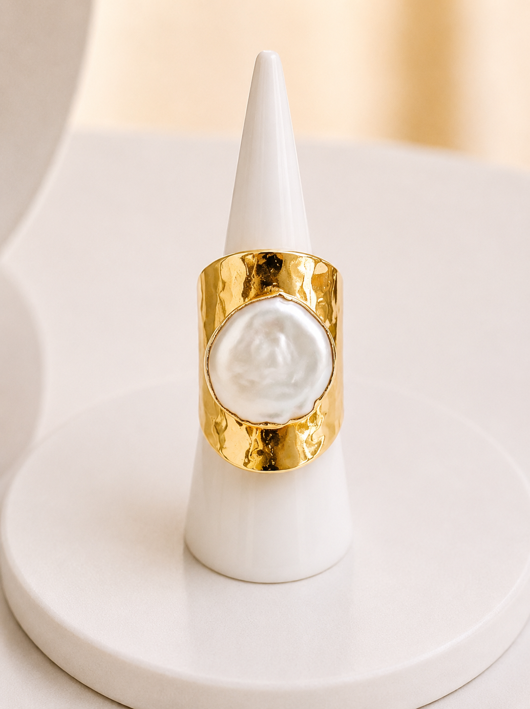 A gold ring with a large, irregular white pearl-like center stone, displayed on a white cone jewelry stand against a soft beige background.