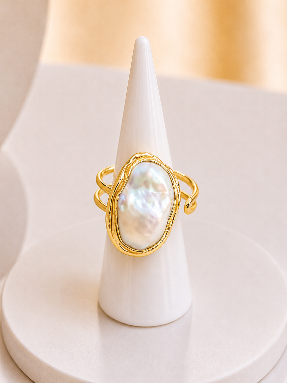 A gold ring with a large, irregular-shaped white baroque pearl, displayed on a white cone jewelry stand against a soft beige background.