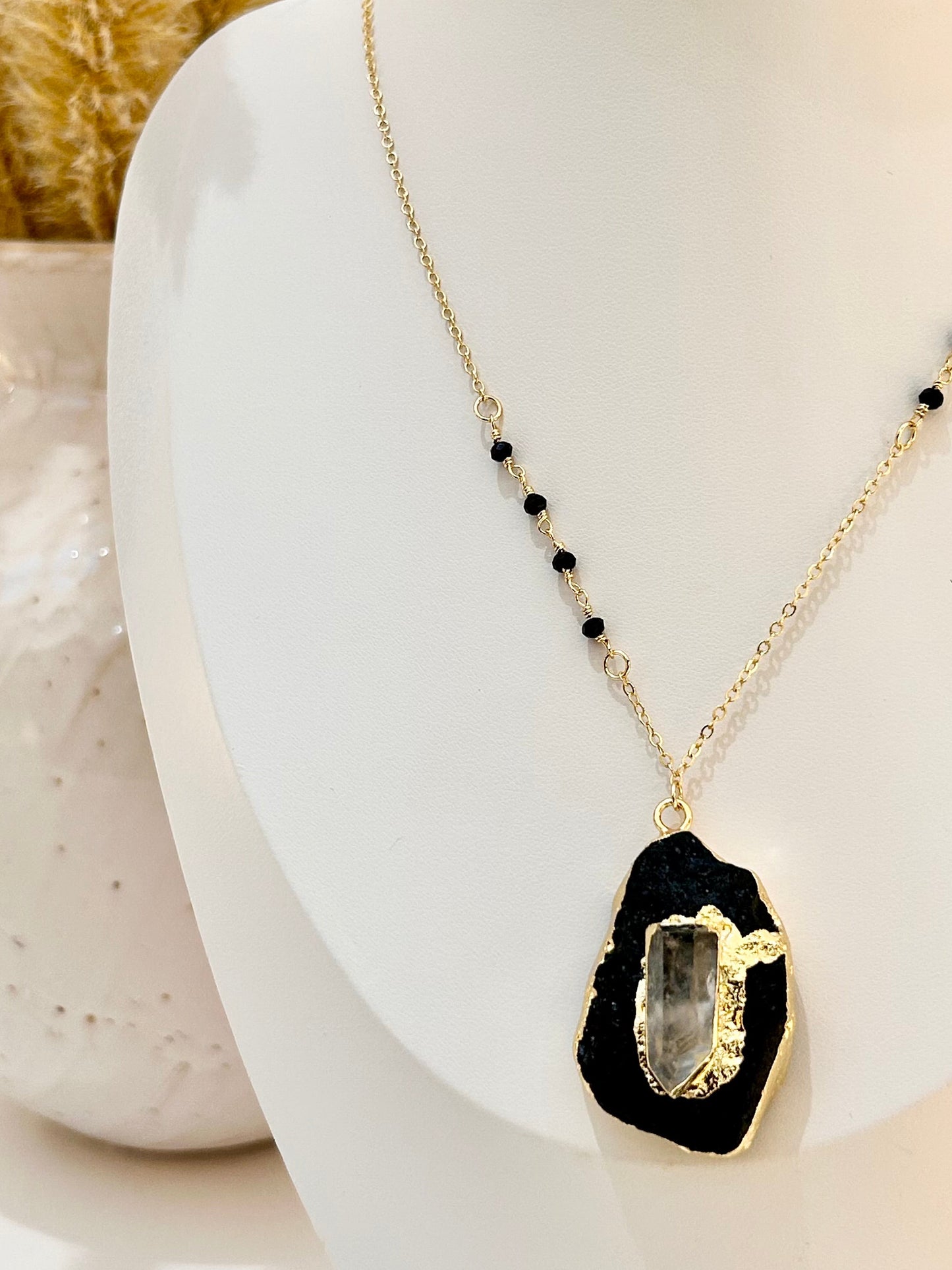 
                  
                    A gold chain necklace with a black agate slice pendant and small black beads, displayed on a white mannequin bust.
                  
                