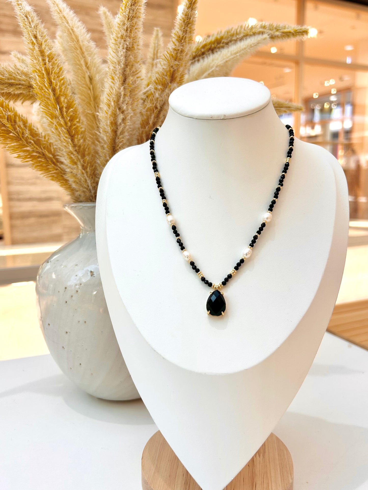 
                  
                    A close-up of a woman wearing a beaded necklace with black obsidian and white pearl beads and a teardrop-shaped black obsidian pendant with gold detailing, paired with a beige lace-trimmed top.
                  
                