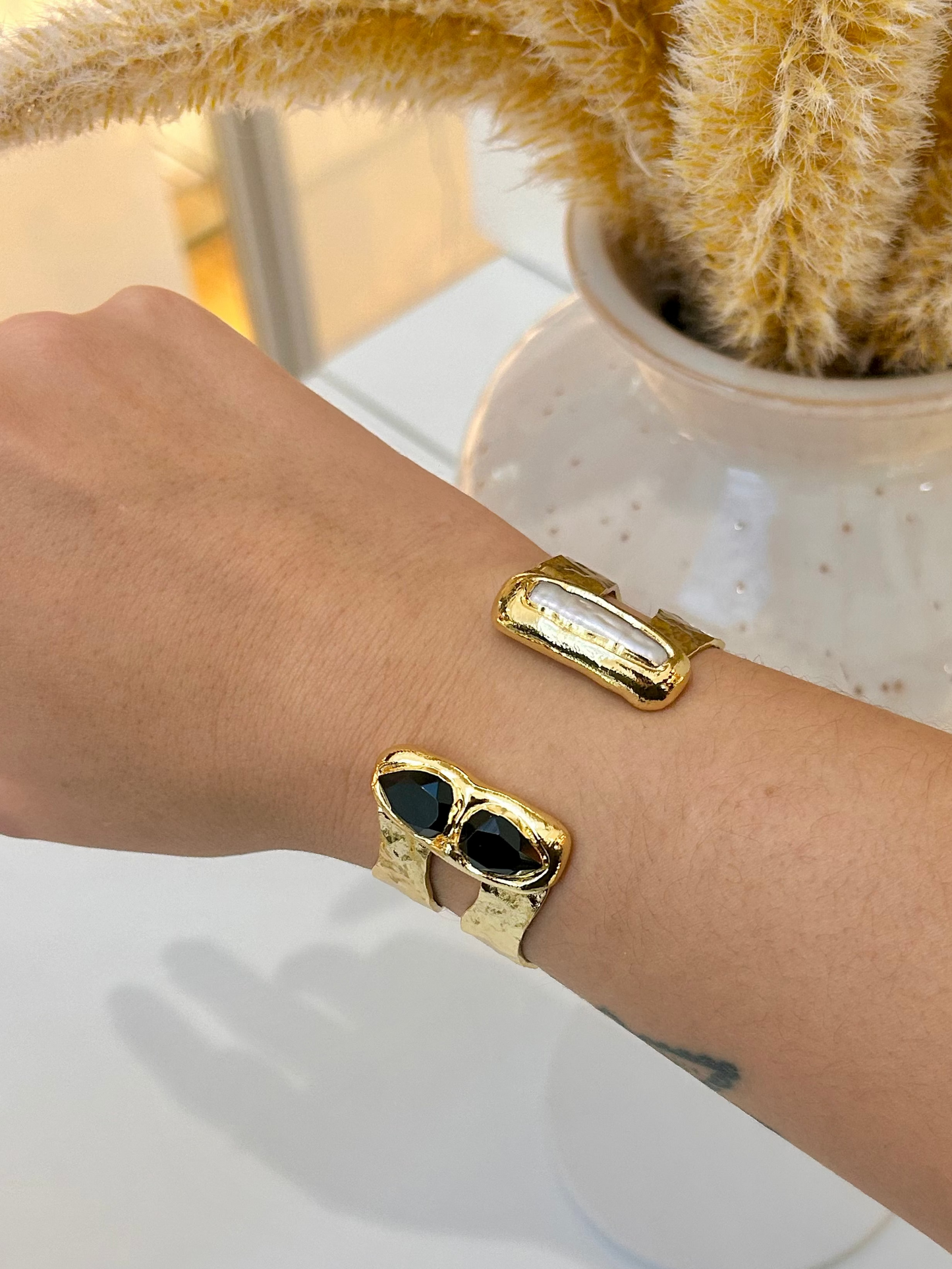 
                  
                    A close-up of a person's forearm wearing gold bangles: one with a freshwater pearls inlay and the other with black obsidian inlay, set against a bright indoor background with a white speckled vase and dried yellow grass.
                  
                