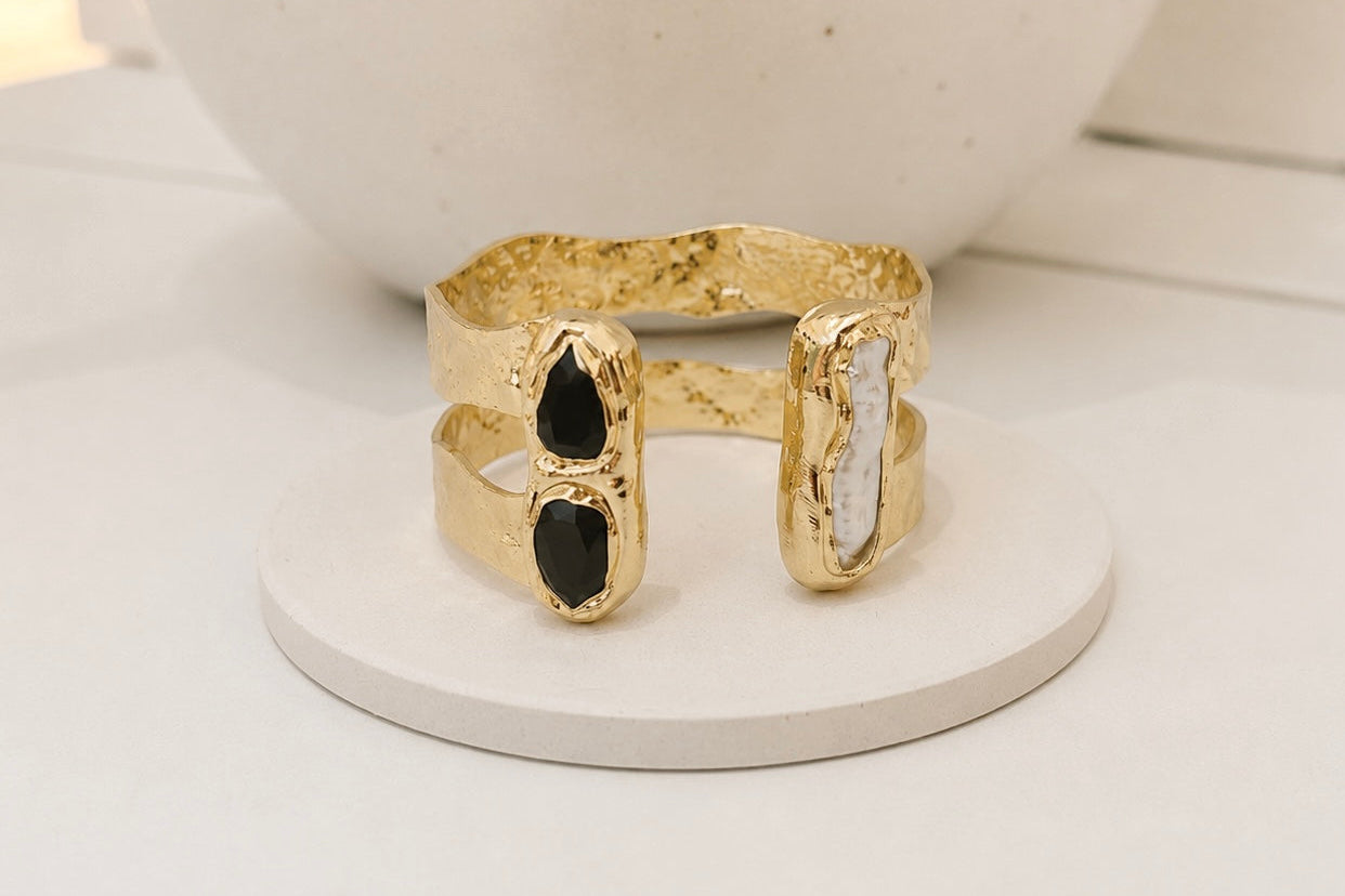 
                  
                    Gold-plated double-band open cuff bracelet featuring two black obsidian stones and a long freshwater pearl in organic settings, displayed on a white jewellery stand.
                  
                