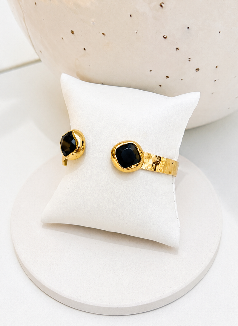 
                  
                    A pair of gold rings with round black gemstones displayed on a small white pillow, set against a neutral white background with a textured ceramic vase.
                  
                