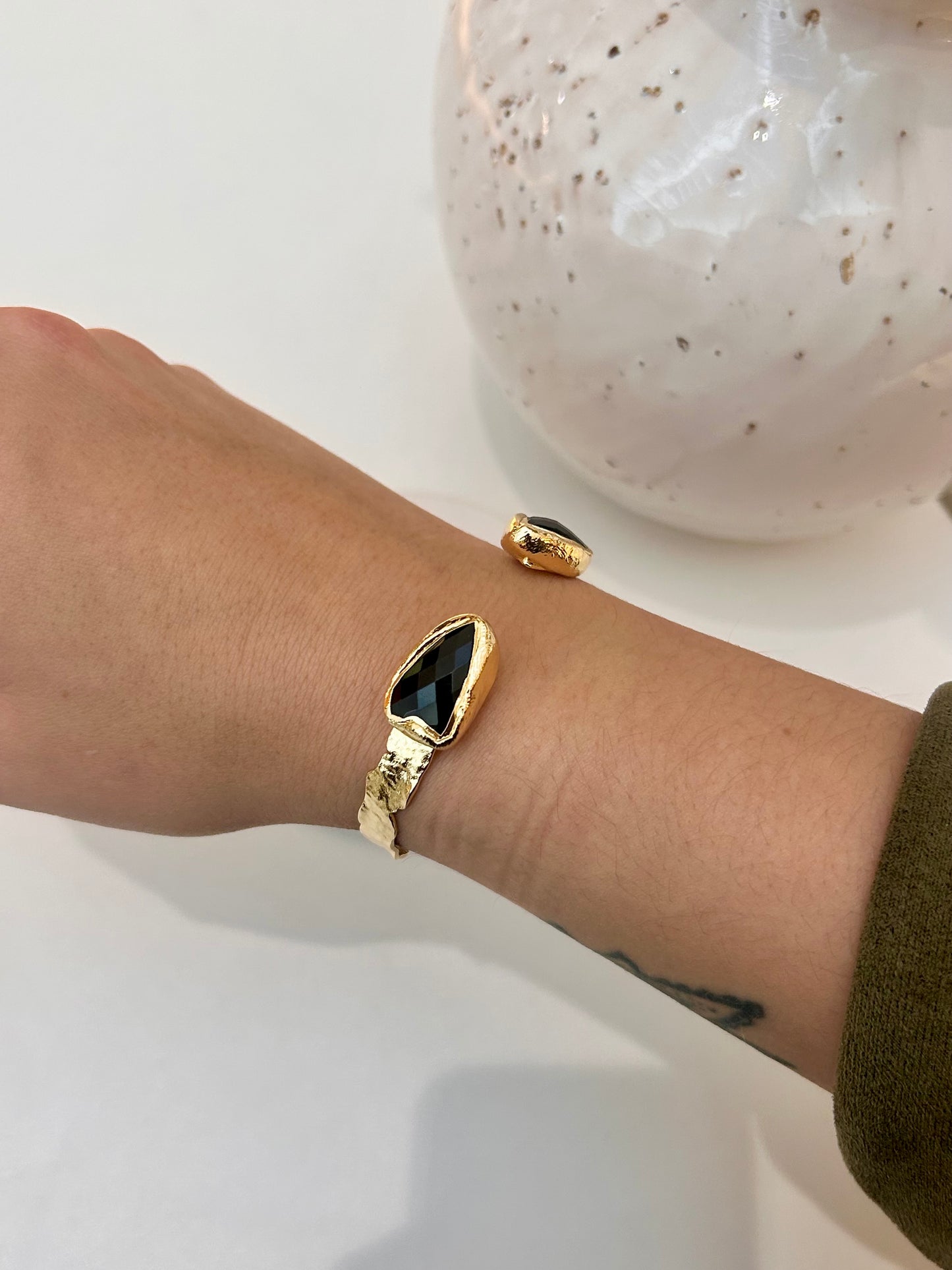 A gold cuff bangle with a teardrop-shaped black obsidian stone, worn on a person's wrist next to a speckled white ceramic vase.