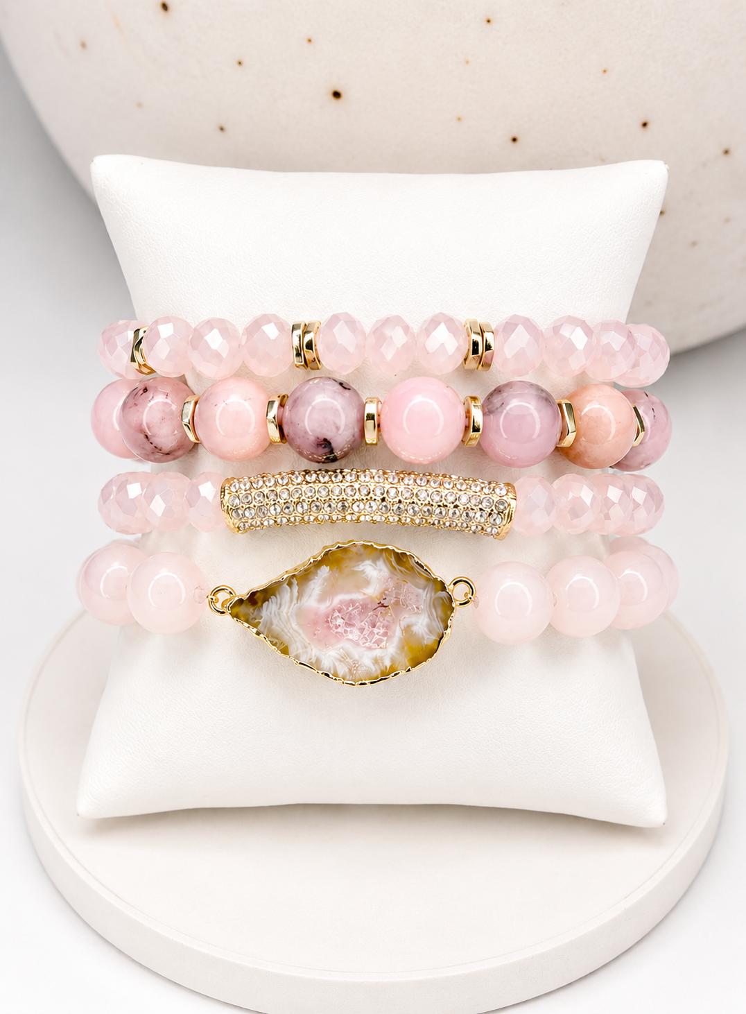 Mixed pink rose quartz beaded bracelets with gold accents and a geode charm, displayed on a white cushion stand.