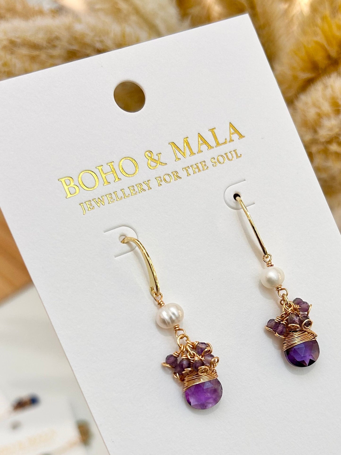 
                  
                    Pair of gold and purple earrings on a BOHO & MALA branded card.
                  
                