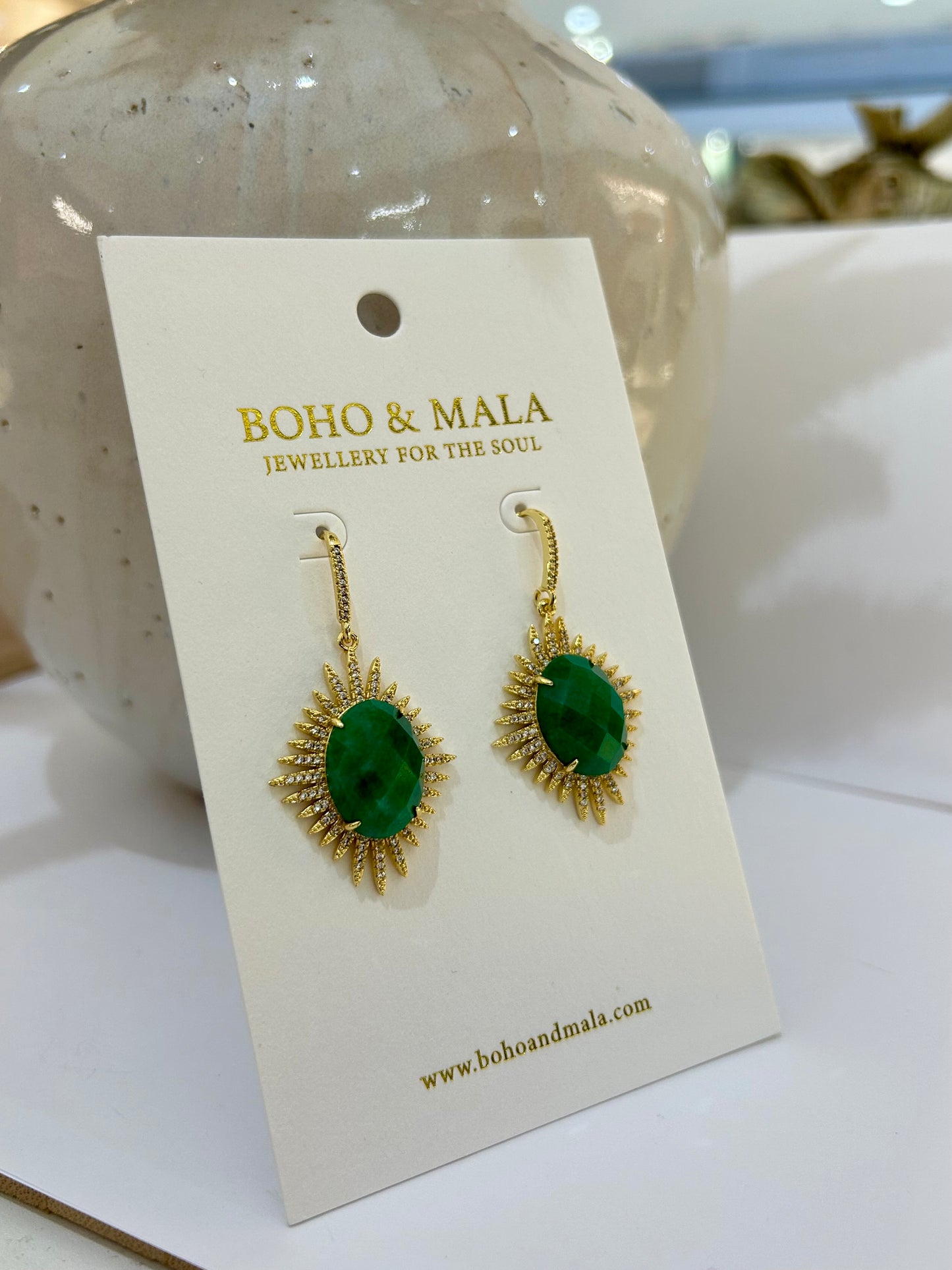 
                  
                    Gold earrings with green gemstones on a BOHO & MALA branded card.
                  
                