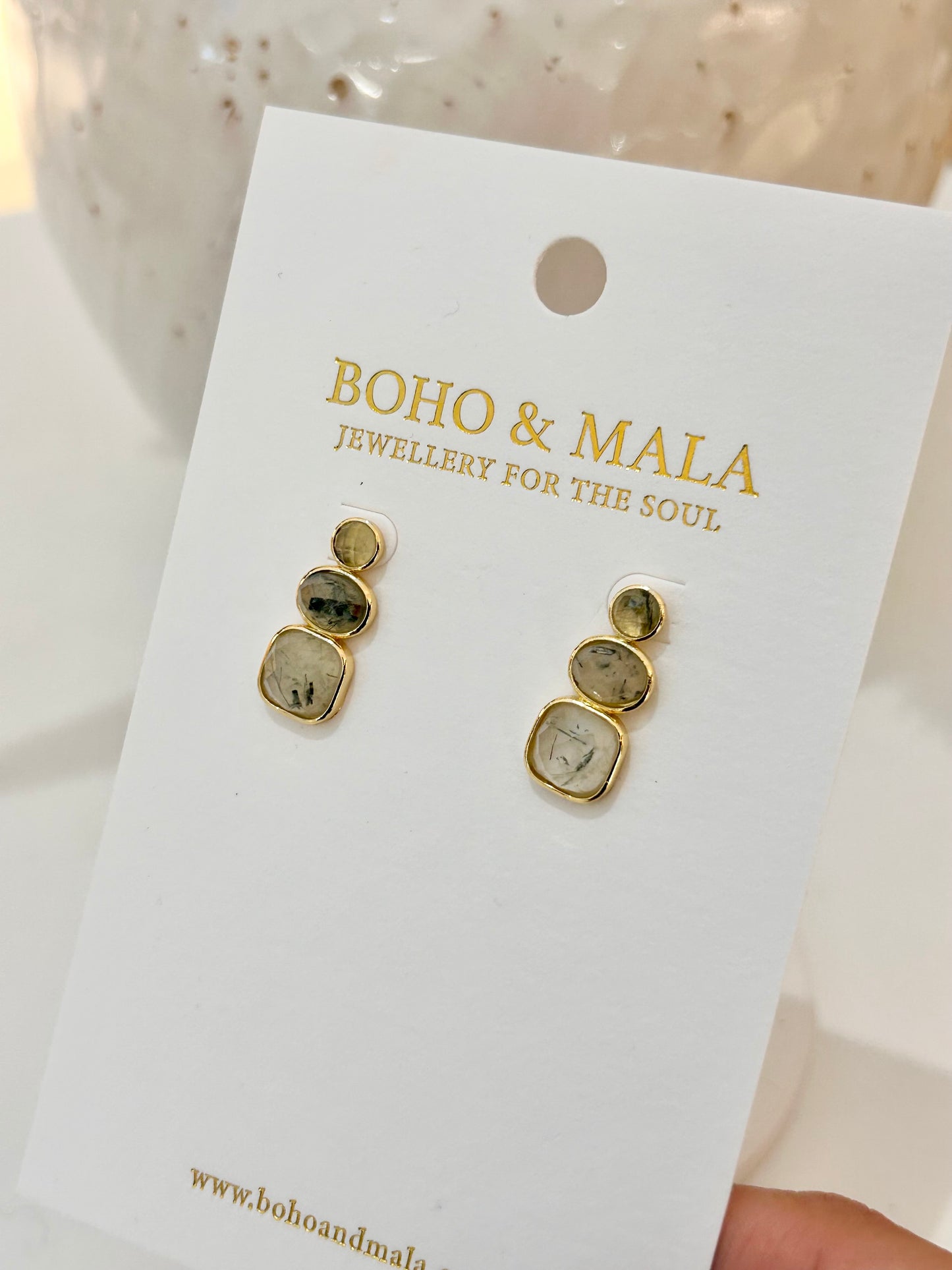 
                  
                    Gold earrings on a 'Boho & Mala' branded card with a blurred background
                  
                