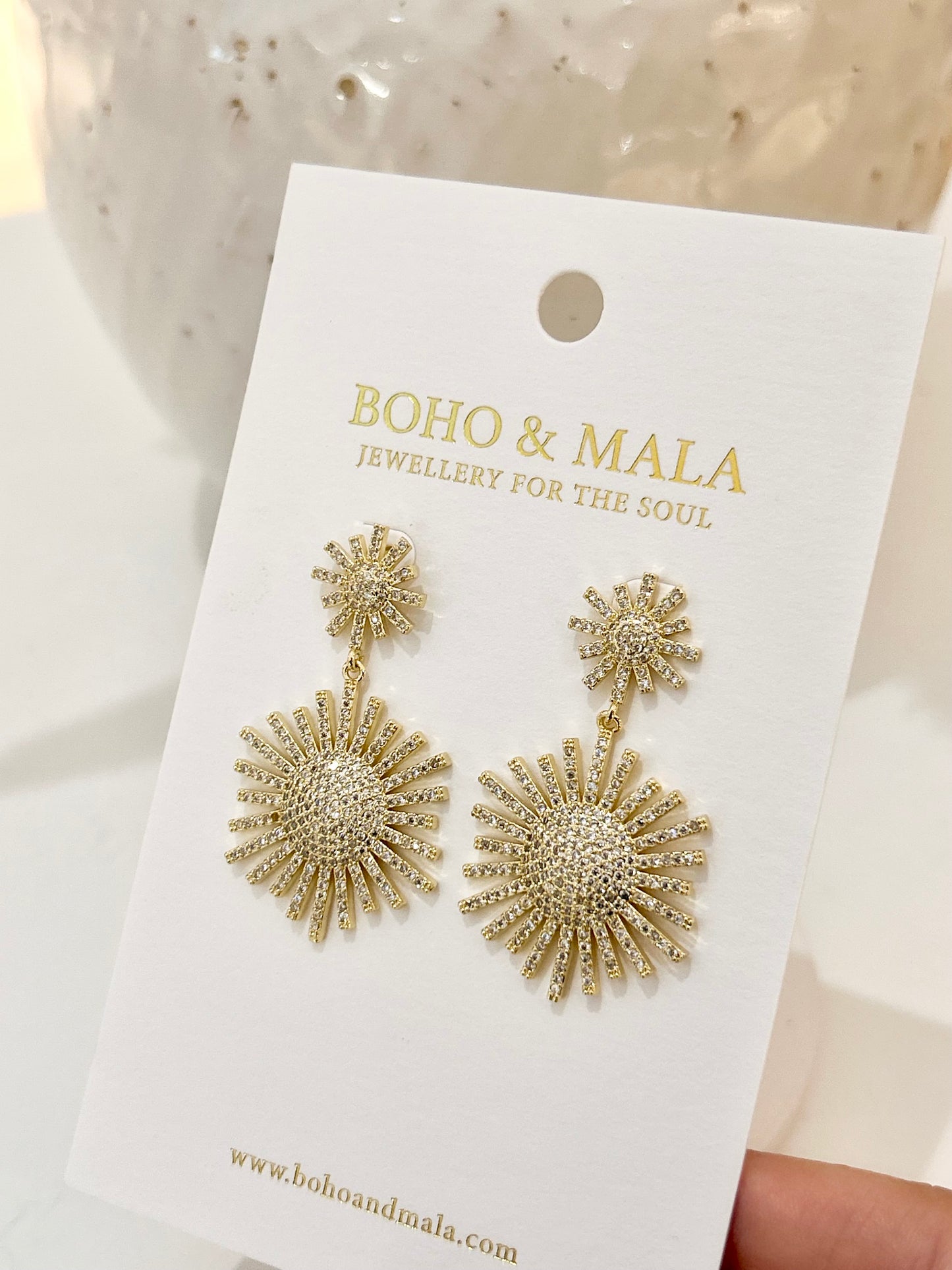 
                  
                    Gold sunburst earrings on a 'Boho & Mala' branded card.
                  
                