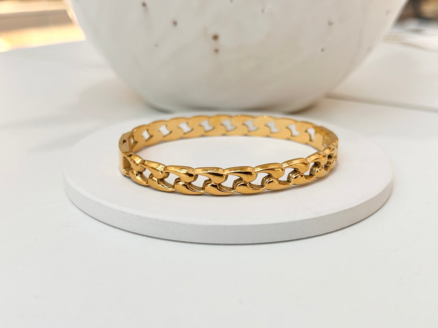 
                  
                    A gold-toned chunky curb chain bangle bracelet displayed on a round white coaster, with a blurred white ceramic bowl in the background.
                  
                