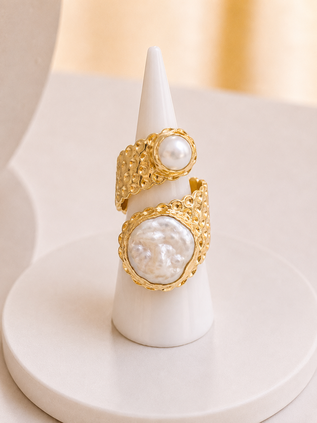 Two gold rings with large baroque pearls displayed on a white cone stand against a soft beige background.