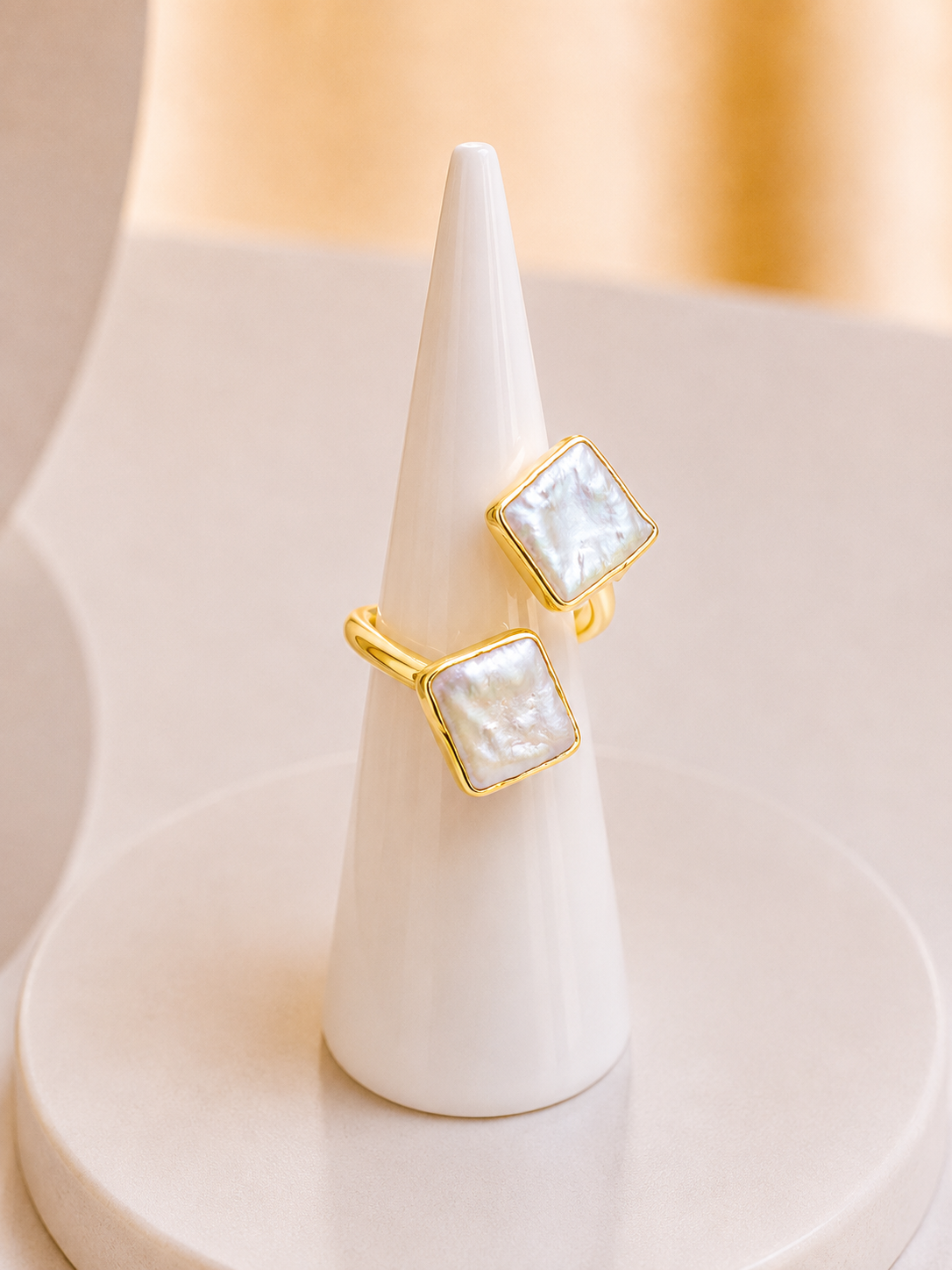 Two gold rings with square white mother-of-pearl inlays displayed on a white cone stand against a soft beige background.