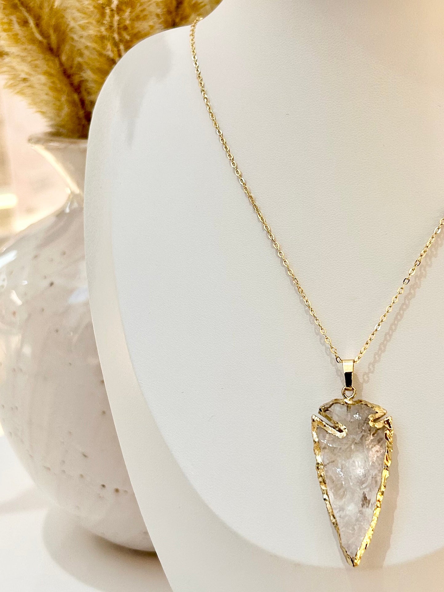 
                  
                    A gold chain necklace with a large, textured clear crystal pendant, displayed on a white mannequin bust with dried pampas grass in the background.
                  
                