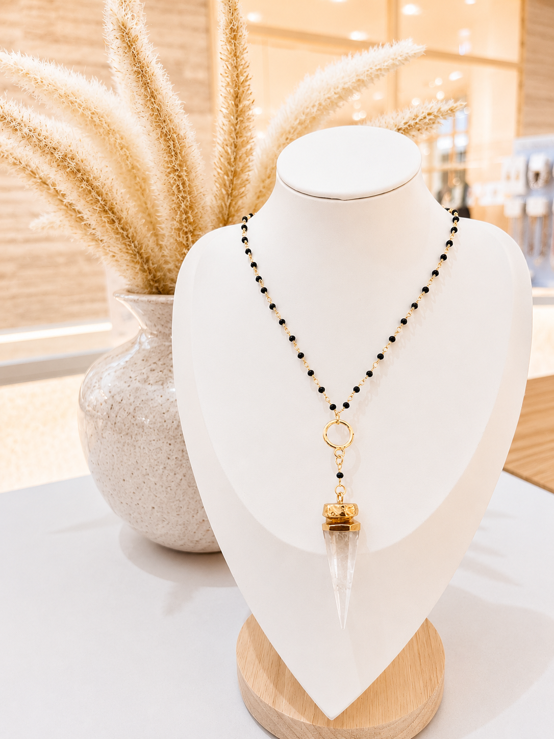 
                  
                    A beaded necklace with a gold-toned ring and a clear pointed pendant, displayed on a white jewelry bust in a softly lit interior setting with dried grass decor in the background.
                  
                