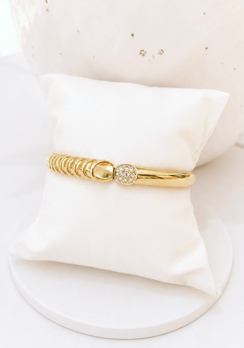 A gold bangle bracelet with a textured segmented design and a round bezel-set crystal accent, displayed on a white satin pillow.