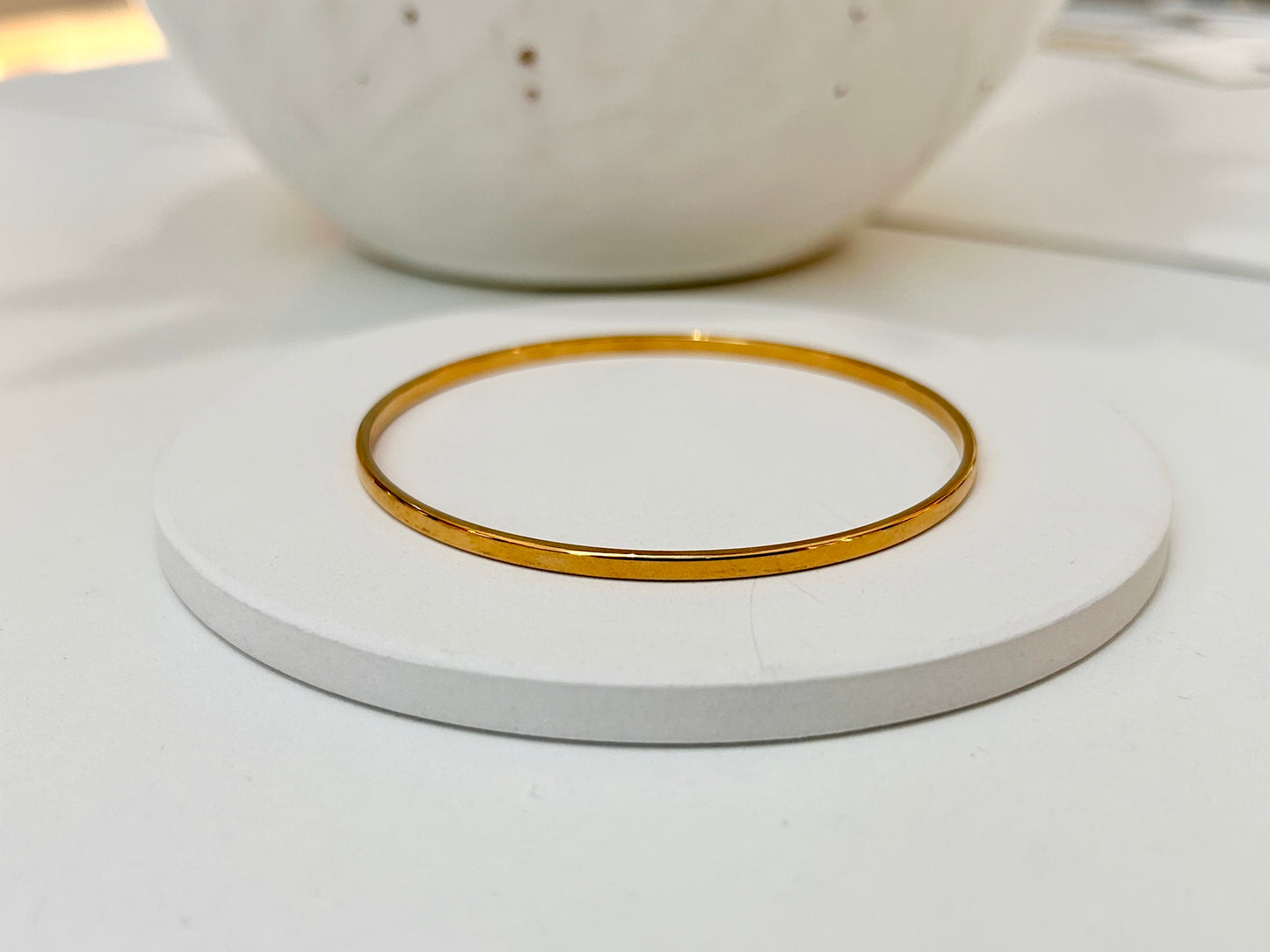 
                  
                    A thin, polished gold bangle bracelet displayed on a round white coaster, with a blurred white ceramic bowl in the background.
                  
                