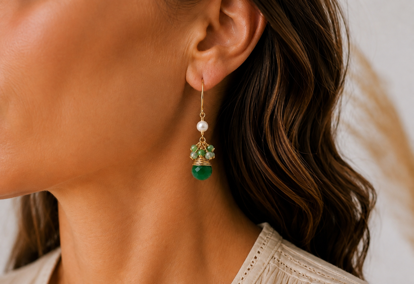 A close-up lifestyle shot of a woman wearing a gold drop earring with a white freshwater pearl, green jade gemstone beads, and small clear crystals.