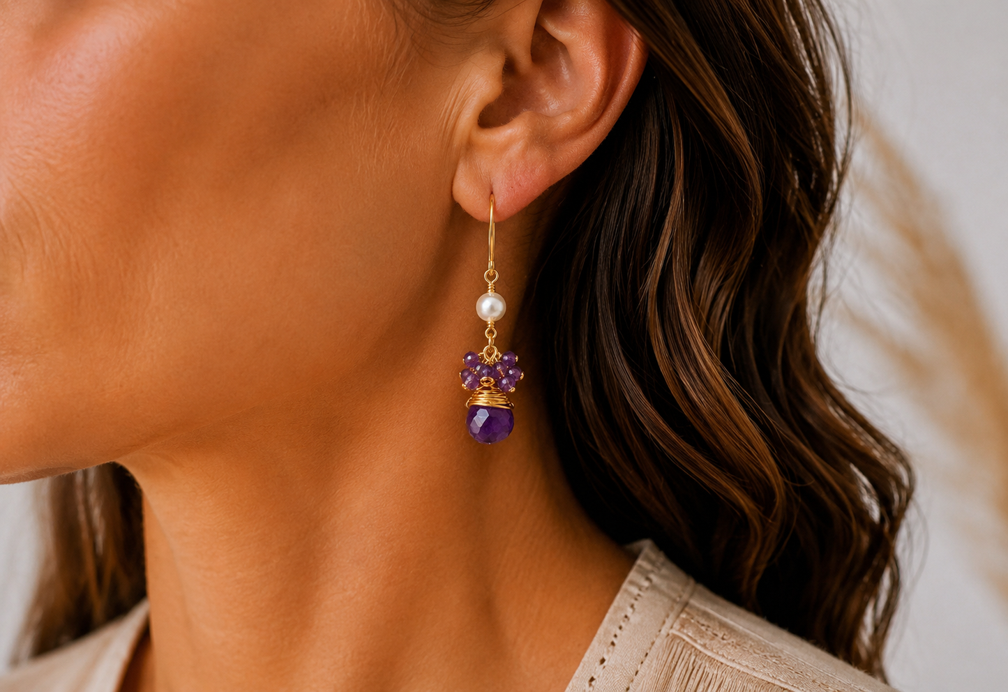 A close-up of a woman's ear wearing a dangling earring with a gold hook, a white freshwater pearl, purple amethyst crystals, and a faceted purple amethyst gemstone.