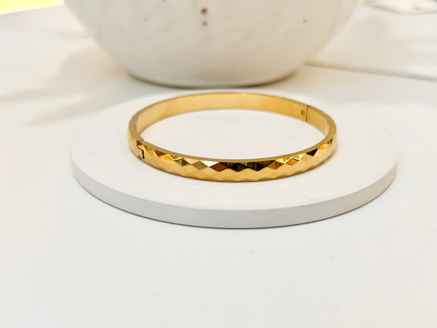 
                  
                    A gold bangle bracelet with a textured, diamond-cut pattern, displayed on a round white coaster.
                  
                